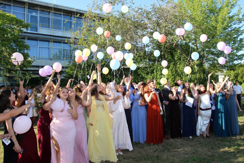 Absolventinnen und Absolventen lassen im Schulgarten bunte Luftballons steigen; festliche Kleider und Anzüge. Im Hintergrund Schulgebäude und grüne Bäume, sonniger Abend.