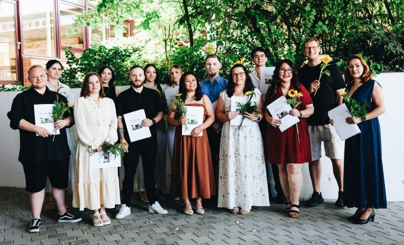 Pflegefachfrauen/-männer/-personen mit ihrem Zeugnis, 2. von rechts: Klassenlehrer Dirk Urbschat-Bau Gruppenfoto vor der Schule: lächelnde Absolventinnen und Absolventen halten Urkunden und Blumen in den Händen; festliche Kleidung, Sommerlicht, grüne Büsche im Hintergrund. 2. von rechts: Klassenlehrer Dirk Urbschat-Bau