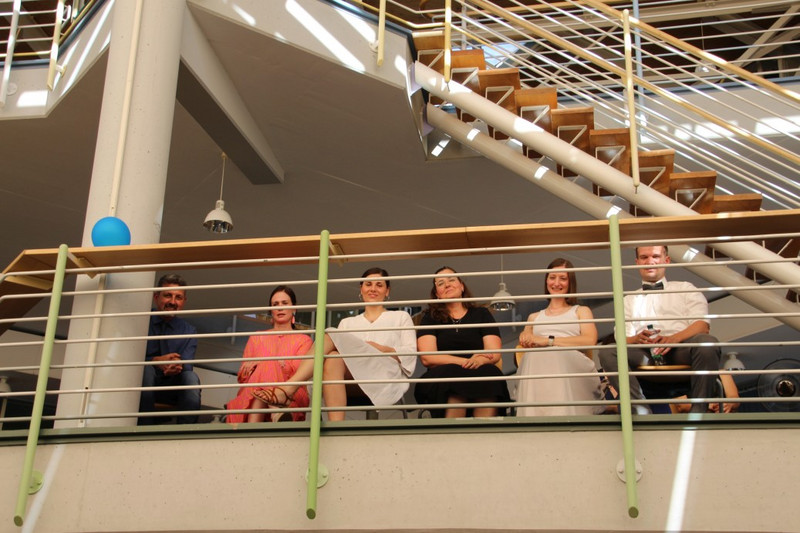 Lehrkräfte sitzen auf der Galerie im Schulatrium und blicken auf die Feier unten; neben ihnen verläuft die Treppe, am Geländer hängt ein blauer Ballon.