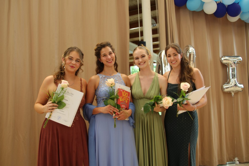 Vier Absolventinnen posieren auf der Bühne in festlichen Kleidern, halten Rosen, Urkunden und ein Buch; im Hintergrund beige Vorhänge, silberne Ballons und blau‑weiße Ballongirlande.