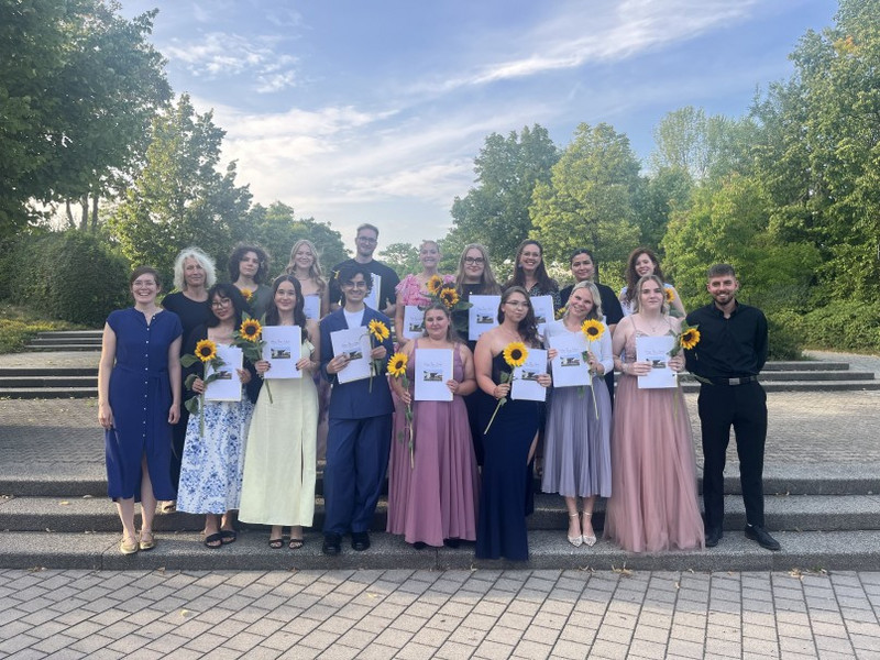 Gruppenfoto auf Steintreppen: Absolventinnen und Absolventen halten Urkunden und Sonnenblumen, festlich gekleidet. Hinter ihnen Bäume und Abendhimmel; weitere Gäste stehen im Hintergrund.