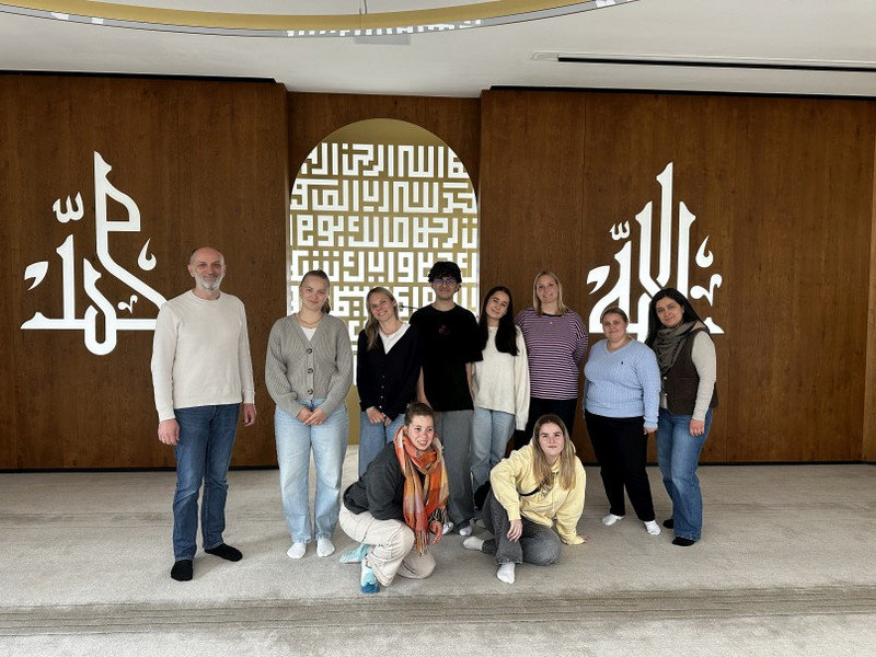 Besamin Limani von der Moscheegemeinde mit Schüler:innen Besamin Limani von der Moscheegemeinde (links) mit Schüler:innen in einem modernen Gebetsraum der Moschee; hölzerne Wände mit großer arabischer Kalligrafie, dekoratives Bogenfenster im Hintergrund.