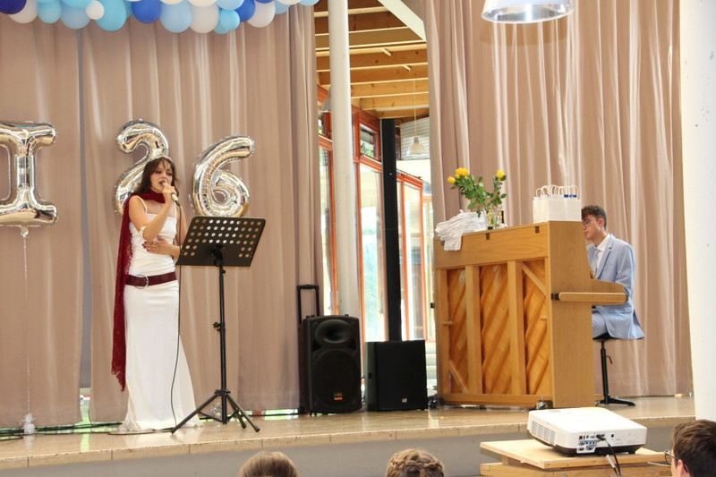 Musikalischer Beitrag bei der Abschlussfeier: Eine Schülerin singt am Mikrofon, ein Schüler begleitet am Klavier. Auf der Bühne Vorhang, Lautsprecher, silberne „26“-Ballons und blau‑weiße Ballongirlande.