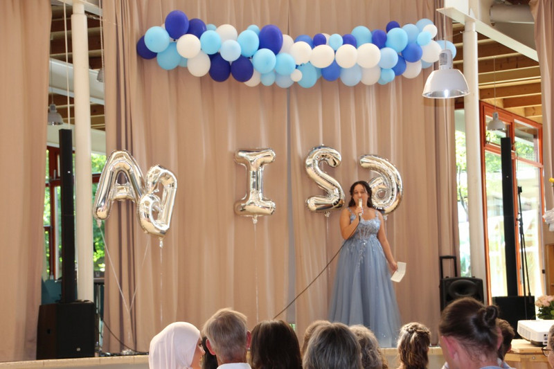 Abschlussfeier im Schulfoyer: Schülerin im blauen Abendkleid moderiert am Mikrofon auf der Bühne; darüber blau‑weiße Ballongirlande, dahinter Vorhang; silberne Ballons formen „ABI 26“; Publikum sitzt davor.