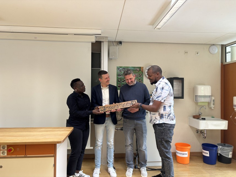 Dr. Albrecht Weber (Mitte rechts) und Daniel Fegert (Mitte links) mit den beiden Gästen aus Kenia im Klassenzimmer der Hilde-Domin-Schule Im Klassenraum überreichen die beiden Gäste aus Kenia (außen) Daniel Fegert (Mitte links) und Dr. Albrecht Weber (Mitte rechts) ein Geschenk: ein Holzschild mit der Aufschrift „Hakuna Matata“. Im Hintergrund Whiteboard, Waschbecken und Mülltrennungstonnen.