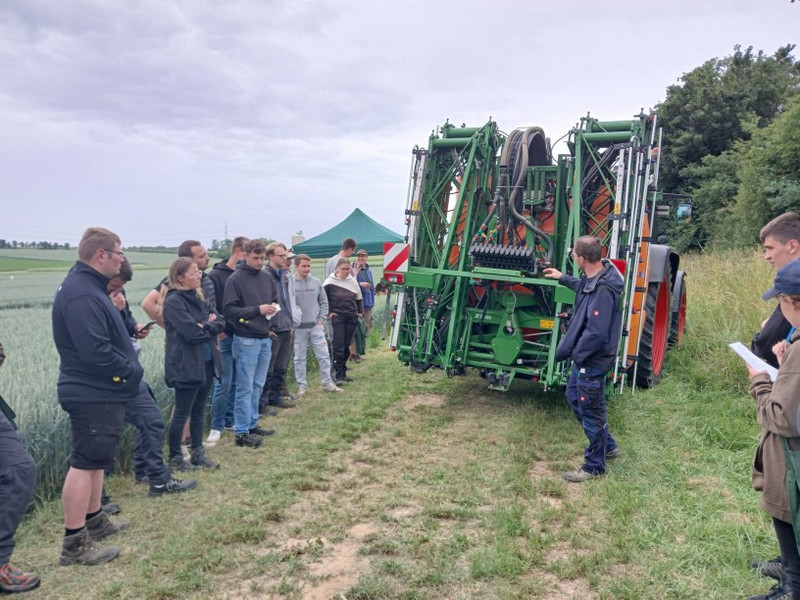 Pflanzenschutzspritze Gruppe Lernender am Feld; eine Person erklärt eine große grüne Feldspritze am Traktor, Zelt im Hintergrund.