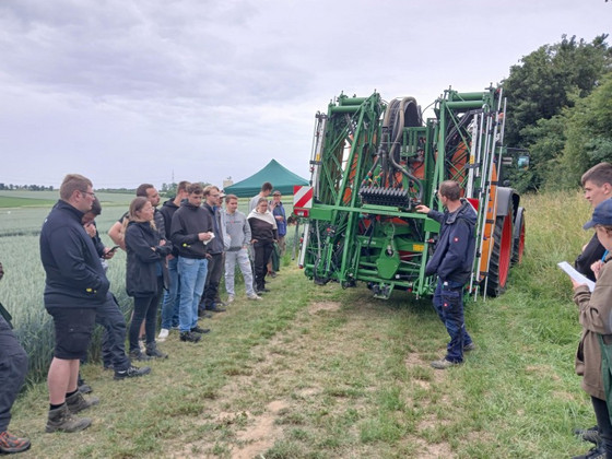 Gruppe Lernender am Feld; eine Person erklärt eine große grüne Feldspritze am Traktor, Zelt im Hintergrund.