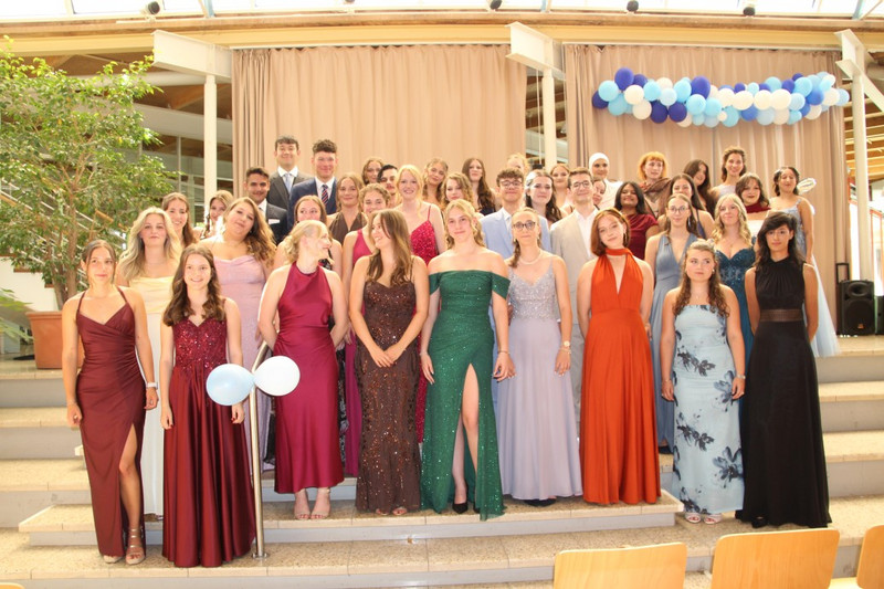 Abiball im Schulfoyer: Schülerinnen und Schüler posieren auf Stufen in festlicher Abendkleidung; hinter ihnen Vorhang und blau‑weiße Ballondeko. Fröhliche Gruppenaufnahme bei der Abschlussfeier.