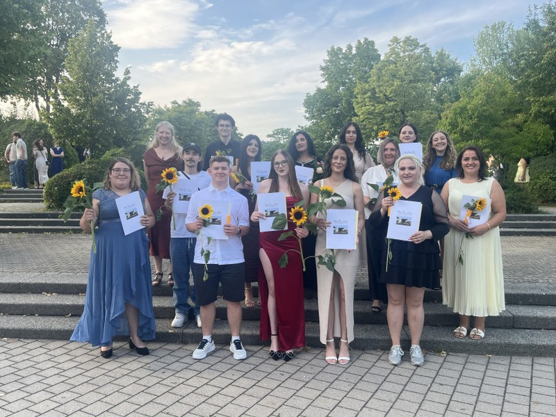 Gruppenfoto im Freien: Erwachsene stehen auf Stufen, halten Urkunden und Sonnenblumen; sommerlicher Abendhimmel, grüne Bäume im Hintergrund. Abschlussfeier der Schule.