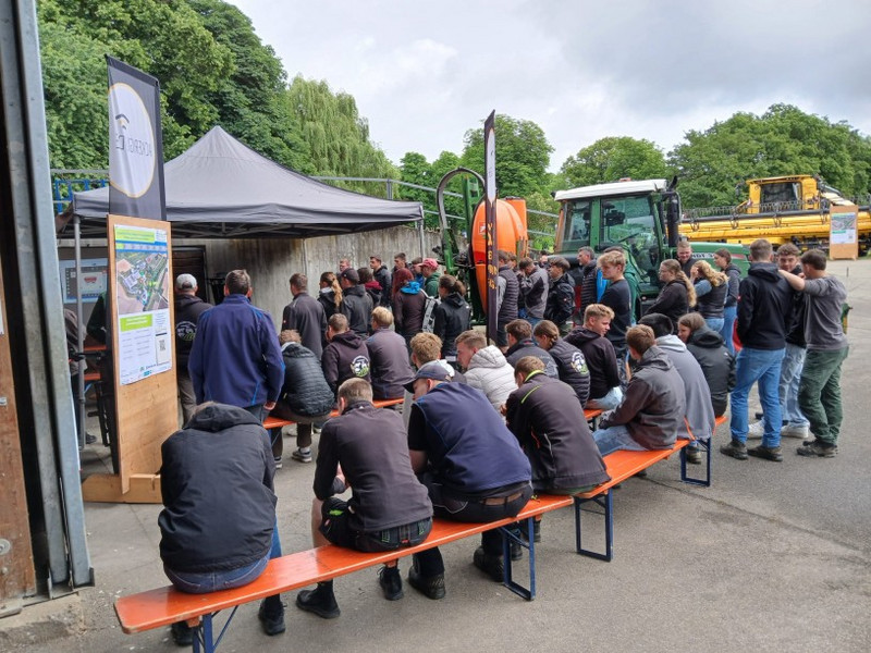 Station 1: Nachrüstlösungen Automatisierung Schülerinnen und Schüler besuchen eine Außenveranstaltung: Unter einem Pavillon erläutern Fachleute Landtechnik; auf Bänken sitzen Gruppen, nebenan stehen Traktor und Mähdrescher.