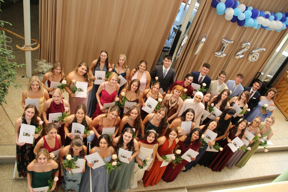 Absolventinnen und Absolventen posieren im Schulatrium, von oben fotografiert: festliche Kleidung, jede und jeder hält Urkunde und Rose. Hinter ihnen Vorhang, blau‑weiße Ballongirlande und silberne Jahrgangsballons.