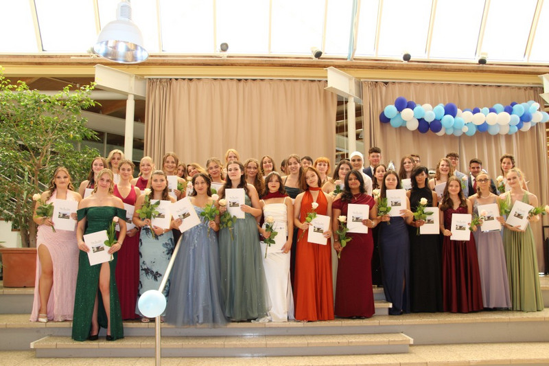 Absolventinnen und Absolventen stehen auf den Stufen im Schulatrium, festlich gekleidet, halten Urkunden und Rosen; hinter ihnen beige Vorhänge, blau‑weiße Ballongirlande und viel Tageslicht.