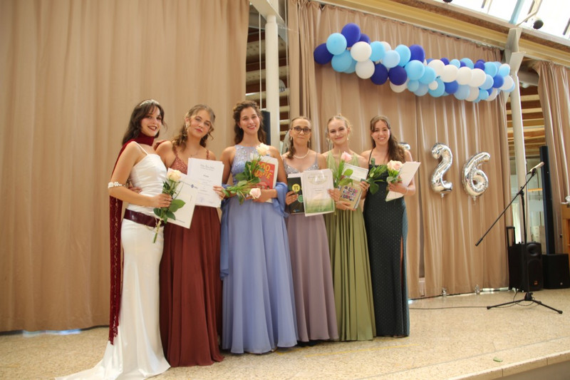 Sechs Absolventinnen posieren auf der Bühnenstufe in festlichen Kleidern, halten Urkunden, Bücher und Rosen. Hinter ihnen beige Vorhänge, blau‑weiße Ballongirlande und silberne Jahrgangsballons „26“, Mikrofonständer rechts.