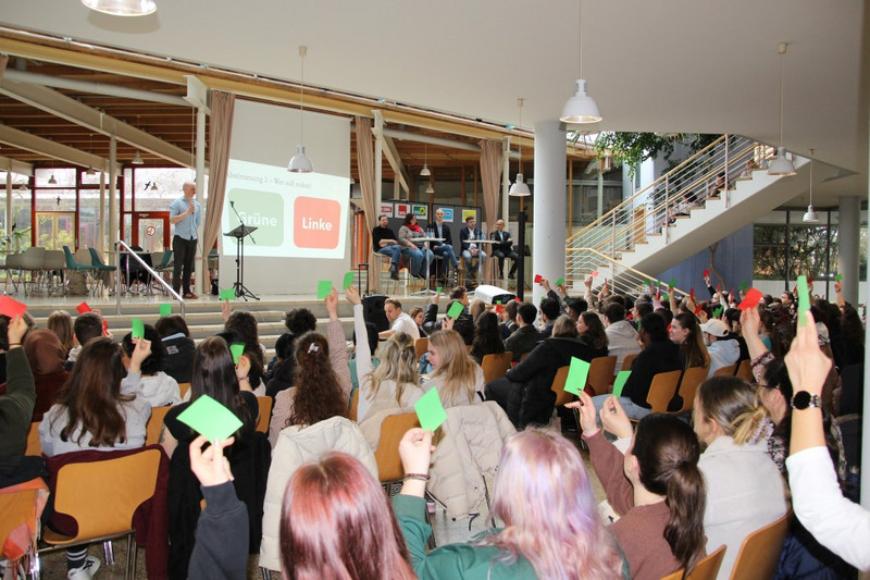 Blick durch das Foyer auf die Bühne, Schülerinnen und Schüler stimmen mit farbigen Zetteln über die Gegner des Rededuells ab