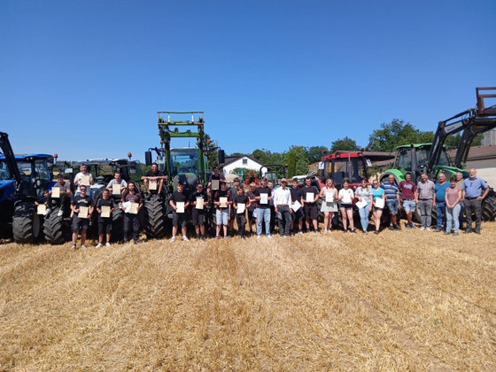 Gruppenfoto auf dem Stoppelfeld: Schülerinnen, Schüler erhalten Zeugnisse mit Lehrkräften; hinten stehen mehrere Traktoren. Sonniger Abschluss eines landwirtschaftlichen Praxistags.