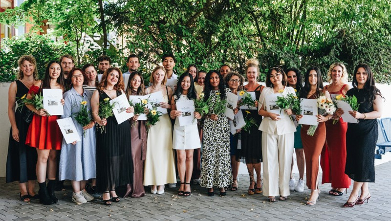 Altenpflegehelfer*innen mit ihren Zeugnissen, links Klassenlehrerin Anna Kleefeld Gruppenfoto von lächelnden Absolventinnen und Absolventen mit Blumensträußen und Urkunden, draußen unter Bäumen; festliche Kleidung; links Klassenlehrerin Anna Kleefeld
