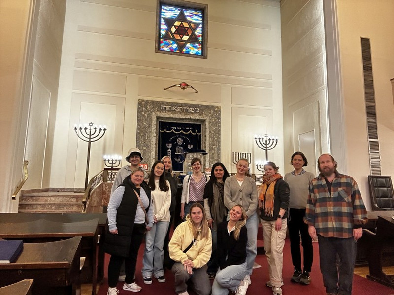 Gruppenfoto der Klasse BKSPIT-PIA 3 mit Lehrkräften. Rechts im Bild Tuviah Christ von der jüdischen Gemeinde Stuttgart. Gruppenfoto von Schülerinnen, Schülern und Lehrkräften in der Stuttgarter Synagoge: vor dem Toraschrein, flankiert von Menora-Leuchtern; oben ein farbiges Fenster mit Davidstern. Rechts Tuviah Christ