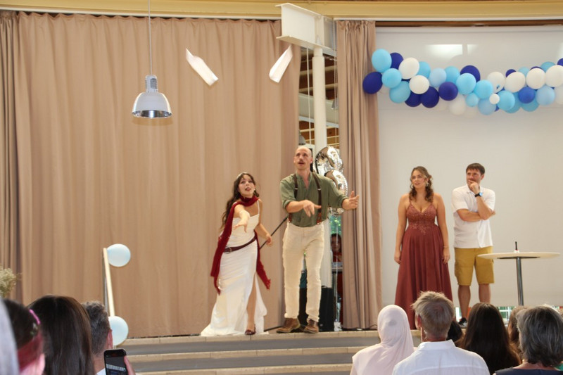 Spiel bei der Abschlussfeier: Schülerin und Lehrer werfen Papierflieger, weitere Personen schauen zu; blau‑weiße Ballongirlande und Vorhang im Hintergrund, Publikum sitzt im Schulatrium.