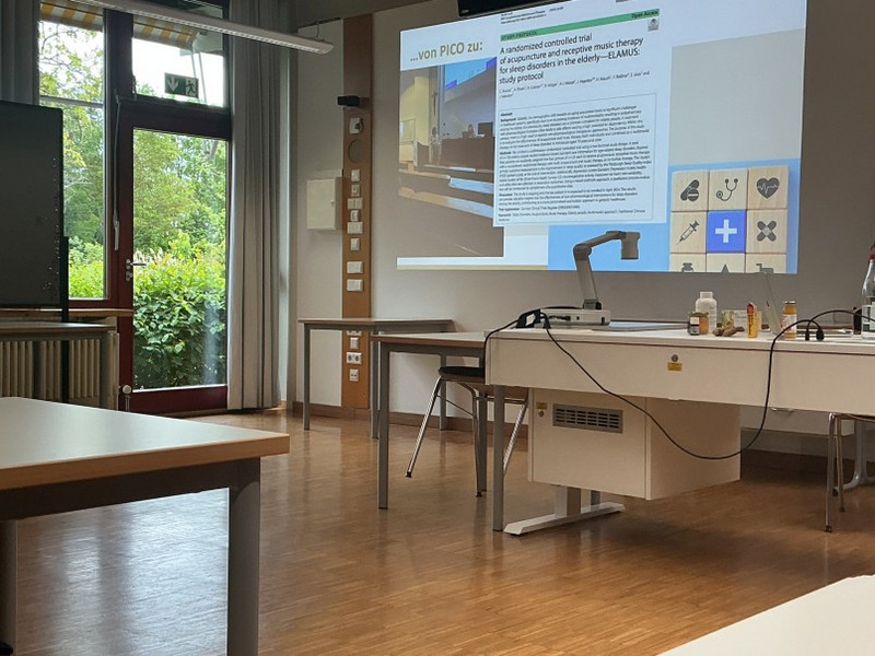 Blick in den Unterrichtsraum Heller Unterrichtsraum: Beamer zeigt Folie zu wissenschaftlicher Studie, Lehrtisch mit Dokumentenkamera und Laborflaschen, leere Schülertische; offene Tür und Fenster mit Blick ins Grüne.