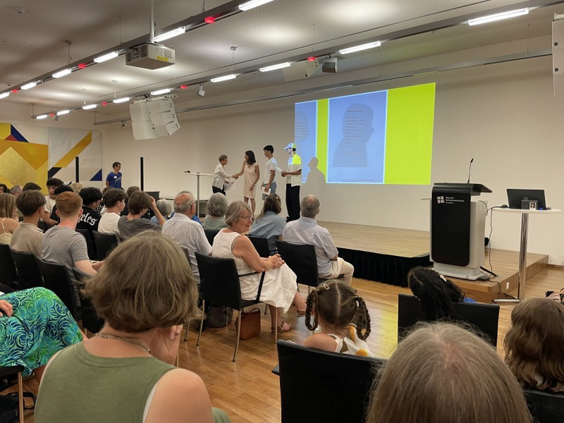 Preisverleihung an Lehrerin Sibylle Schorpp und die beiden Schüler Preisverleihung im Veranstaltungssaal: Publikum sitzt vor Bühne; auf der Leinwand sind Gedichte mit Silhouetten zu sehen. Eine Moderatorin überreicht Lehrerin Sibylle Schorpp und den beiden Schülern Urkunden; Rednerpult rechts.