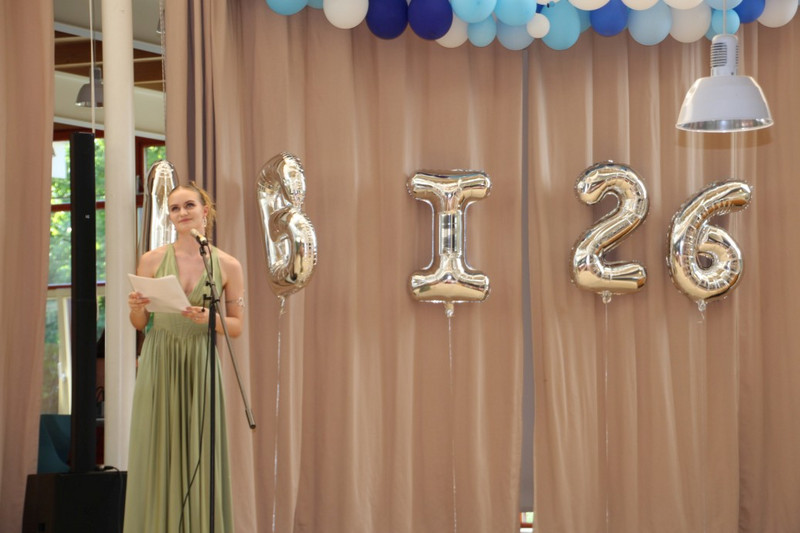 Schülerin im grünen Abendkleid hält eine Rede am Mikrofon auf der Abschlussbühne; hinter ihr beige Vorhänge, blau‑weiße Ballongirlande und silberne Ballons „ABI 26“.