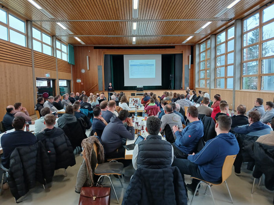 Blick von hinten in der Stadthalle Neubulach auf die Bühne des Milchviehtages; Zuhörer:innen sitzen an langen Tischen 