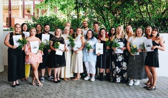 Absolventinnen und Absolventen stehen draußen vor der Schule, halten Urkunden und Blumen, tragen festliche Sommerkleidung; grüne Bäume und Fenster im Hintergrund.