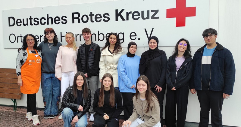 Gruppenfoto der Klasse 1BKSP mit Frau Koebe vom Tafelladen (links im Bild mit Schürze) vor dem Logo des Deutschen Roten Kreuzes