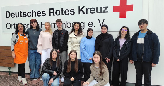 Gruppenfoto der Klasse 1BKSP mit Frau Koebe vom Tafelladen (links im Bild mit Schürze) vor dem Logo des Deutschen Roten Kreuzes