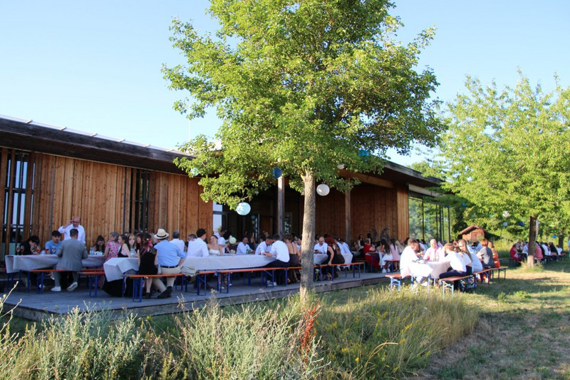Sommerlicher Abschluss des Abiballs im Freien: Viele Gäste sitzen an langen, weiß gedeckten Bänken vor einem Holzpavillon; zwischen Bäumen hängen Dekolichter, warmes Abendlicht.
