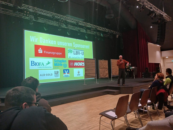 Teilnehmer:innen blicken auf die Vortragsbühne auf dem Junglandwirtekongress in Blaubeuren, ein Vortragender steht auf der Bühne