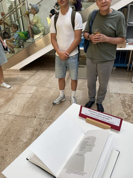 Ausstellung im Foyer: Auf einem Tisch liegt ein geöffnetes Gedicht‑Buch mit Silhouettenporträt; daneben stehen zwei Personen. Im Hintergrund Treppe, sitzende Kinder und helles Tageslicht.