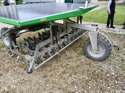 Station 3: Robotics: Aussaat und Pflege Solarbetriebener Landwirtschaftsroboter mit großem Solarpanel und rotierenden Hackwerkzeugen steht auf Kies neben einer Wiese; im Hintergrund stehen Personen, vermutlich bei einer Vorführung.