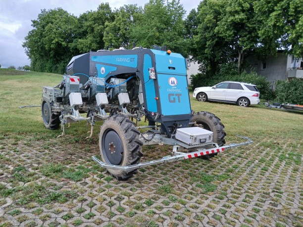 Station 5: Robotics: Hacken Blauer Feldroboter „Farming GT“ mit großen Rädern und Sensorbügeln steht auf einer Wiese; Anbaugeräte sind sichtbar. Im Hintergrund Bäume und ein geparkter weißer Wagen.