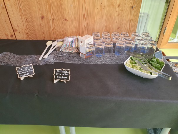 Alkoholfreier Cocktail zum selber mixen Getränkestation auf schwarzem Tisch: viele Gläser, bunte Trinkhalme, Löffel, Schilder „Erfrischung – zum Selbermischen“; Schüssel mit Limetten und Minze. Holzverkleidete Wand, grüner Boden.