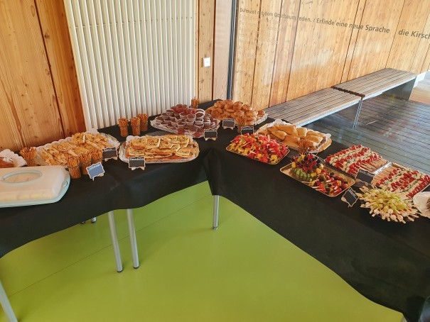 Buffet Buffettische im Neubau mit schwarzen Decken, bestückt mit süßen und herzhaften Snacks: belegte Brötchen, Gebäck, Muffins, Obstspieße und Salzstangen. Holzverkleidete Wand, grüne Bodenfläche, Sitzbank.
