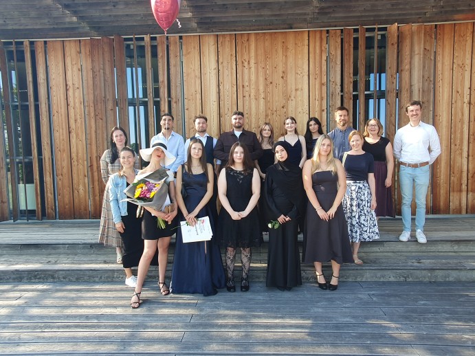 Klasse 2BFP2 mit ihren Lehrkräften Abschlussgruppe vor der Holzfassade der Schule: Schülerinnen und Schüler sowie Lehrkräfte in festlicher Kleidung auf Holzstufen; Blumenstrauß, Urkunden und ein roter Herzballon, sonniger Nachmittag.