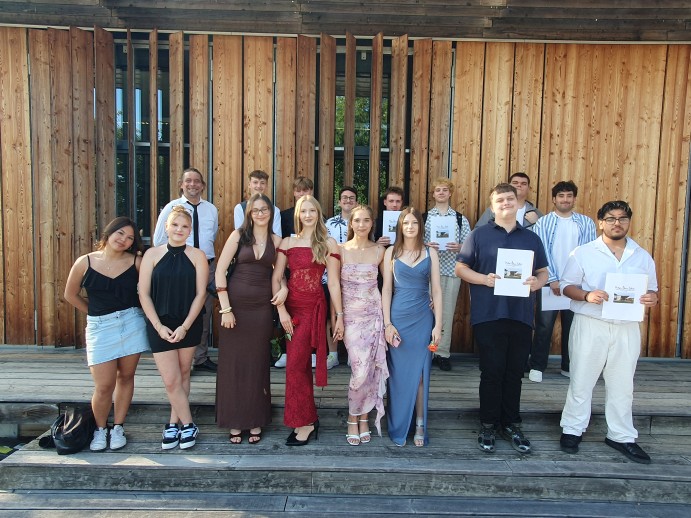 Glückliche und erleichterte Gesichter bei den Absolventinnen und Absolventen der Zweijährigen Berufsfachschule (Klasse 2BFH 2 mit Klassenlehrer Christian Zoller) Gruppe von Schülerinnen und Schülern in festlicher Kleidung posiert draußen vor einer Holzfassade; einige halten Urkunden. Hintere Reihe links: Klassenlehrer Christian Zoller.